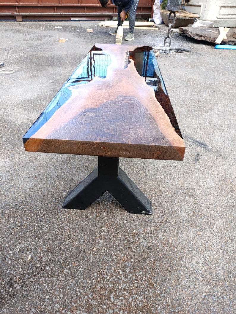 May include: A large, rectangular wooden table with a live edge and a glossy, reflective surface. The table is supported by a black, angular metal base. The wood grain is visible, showcasing natural patterns.