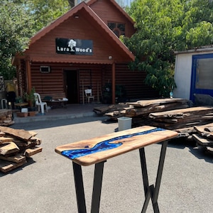 Op de afbeelding: Een houten consoletafel met een rivier van blauwe hars. Het tafelblad is gemaakt van lichtgekleurd hout met een donkerblauwe harstroom. De tafel heeft zwarte metalen poten. De achtergrond toont een houten gebouw met de tekst "Lara Wood's".