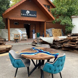 Könnte beinhalten: Ein runder Holztisch mit einem blauen Harzfluss-Design, umgeben von vier tealfarbenen Samtstühlen mit schwarzen Beinen. Der Tisch steht vor einem Holzgebäude mit dem Schild "Lara Wood's".