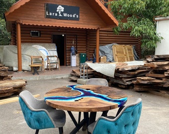 May include: A round wooden table with a blue resin river design, surrounded by four teal velvet chairs with black legs. The table is in front of a wooden building with the sign "Lara Wood's".