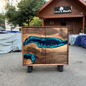 May include: A wooden cabinet with two doors, featuring a blue resin river design. The cabinet is made of dark wood with a natural grain pattern and sits on black feet. The cabinet is displayed outdoors.