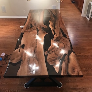 Könnte beinhalten: Ein großer, rechteckiger Esstisch aus Holz mit einzigartigem Design. Die Tischplatte weist ein natürliches Holzmaserungsmuster mit dunklen Harzakzenten auf. Der Tisch wird von einem schwarzen Metallsockel getragen. Der Tisch ist etwa 274 cm lang.