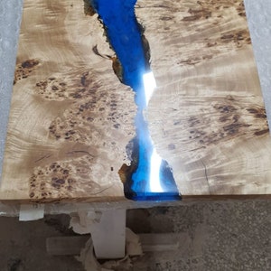 May include: A rectangular wooden table with a river of blue epoxy resin running through the center. The wood has a light brown color with dark brown knots and patterns. The table is supported by a white base.