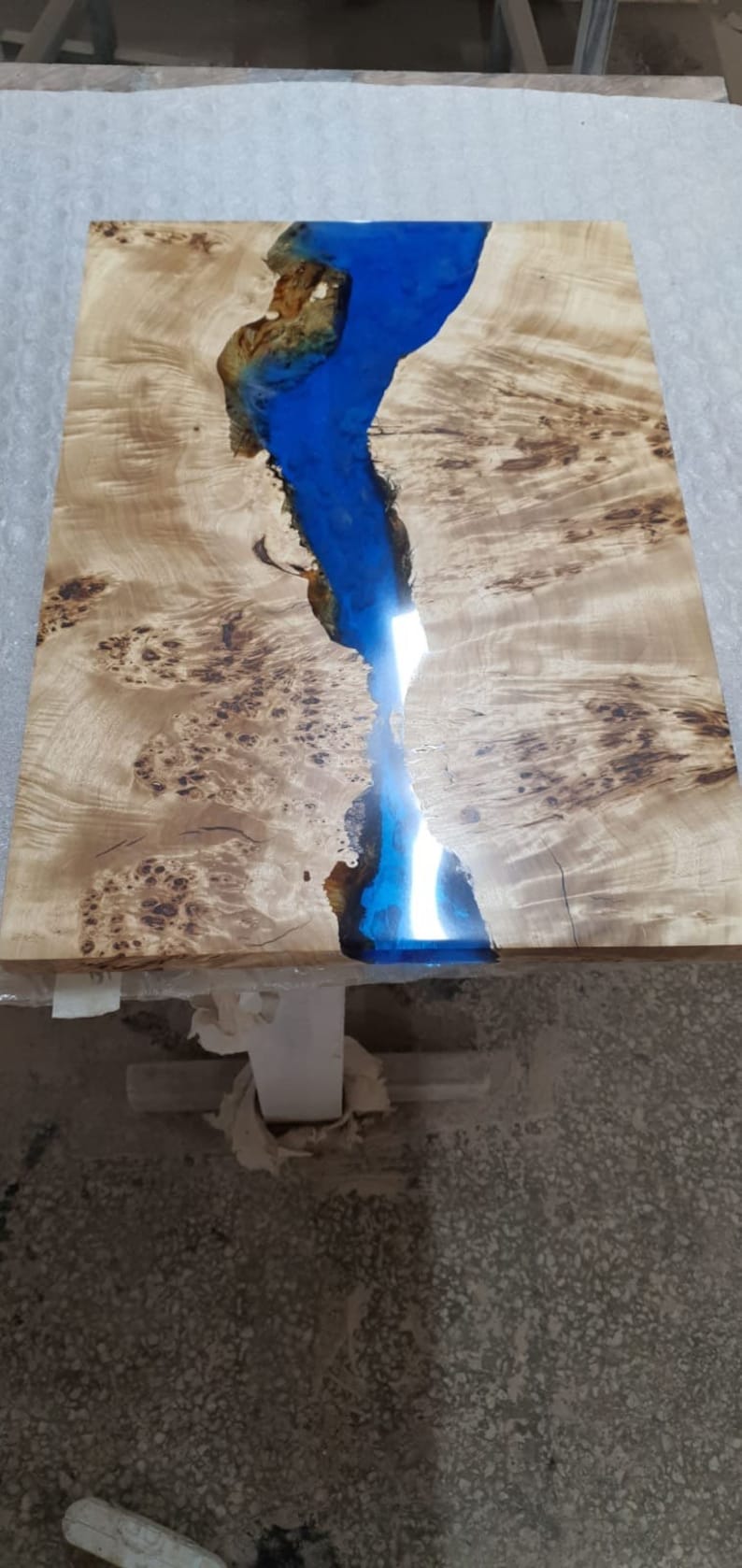 May include: A rectangular wooden table with a river of blue epoxy resin running through the center. The wood has a light brown color with dark brown knots and patterns. The table is supported by a white base.