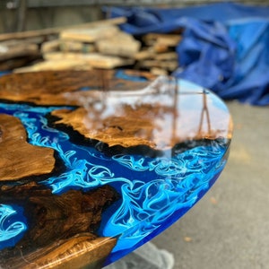 Könnte beinhalten: Nahaufnahme eines handgefertigten runden Tisches mit einzigartigem Design. Der Tisch kombiniert Naturholz und leuchtend blaue Kunstharz, wodurch ein flussartiger Effekt entsteht. Das Holz hat eine satte braune Farbe, das Harz ein Wirbelmuster.