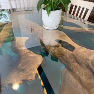 Op de afbeelding: Een op maat gemaakte epoxyhars tafel met een live edge design. De tafel heeft een helder, glanzend oppervlak met een rivier van blauwe hars. Een witte, patroon bloempot met een groene plant staat op de tafel. De tafel is ongeveer 150 cm lang.
