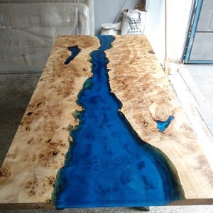 Pode incluir: Mesa de madeira artesanal com design de rio em resina ep&oacute;xi azul. A mesa apresenta um gr&atilde;o de madeira clara com n&oacute;s castanhos escuros e um rio azul brilhante no centro. A mesa &eacute; retangular, com cerca de 180 cm de comprimento.