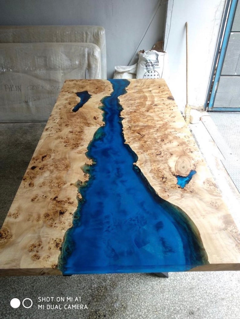 Pode incluir: Mesa de madeira artesanal com design de rio em resina ep&oacute;xi azul. A mesa apresenta um gr&atilde;o de madeira clara com n&oacute;s castanhos escuros e um rio azul brilhante no centro. A mesa &eacute; retangular, com cerca de 180 cm de comprimento.