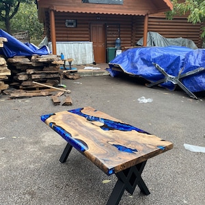 May include: A handcrafted wooden coffee table with a live edge design. The table features a rich, natural wood grain with vibrant blue resin accents. It is supported by black metal legs, creating a modern and unique aesthetic. The table measures approximately 48 inches long.