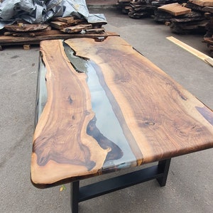 May include: A large, rectangular wooden table with a clear epoxy resin river running through the center. The table has a dark brown finish and sits on a black metal base. The business name "Lara Wood's" is visible in the background.
