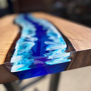 May include: A close-up of a wooden table with a river of blue resin running through the center. The resin is a mix of deep blue and white, creating a wave-like effect. The wood has a natural grain.