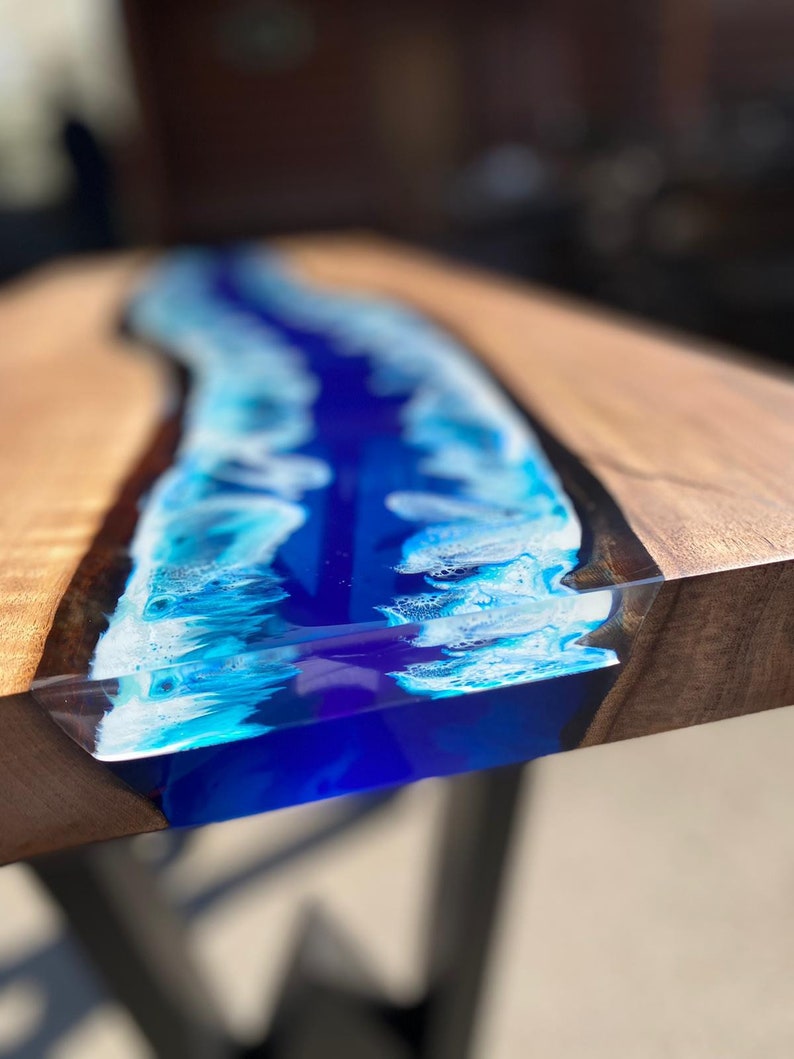 May include: A close-up of a wooden table with a river of blue resin running through the center. The resin is a mix of deep blue and white, creating a wave-like effect. The wood has a natural grain.