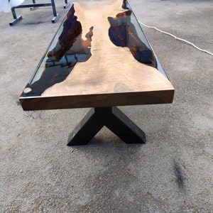 May include: A rectangular wooden coffee table with a dark brown base and a light brown top. The top has a clear resin inlay with dark brown and black accents. The table is supported by a black, angled metal base.