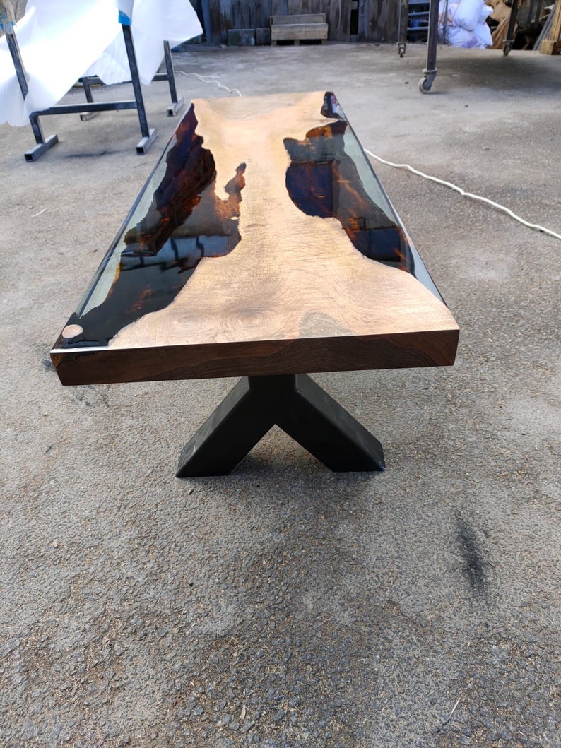 May include: A rectangular wooden coffee table with a dark brown base and a light brown top. The top has a clear resin inlay with dark brown and black accents. The table is supported by a black, angled metal base.