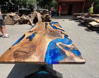 May include: A large, rectangular wooden table with a dark brown finish and a vibrant blue epoxy resin river design. The table is supported by a black metal base. The table is outdoors, with a building and trees in the background.