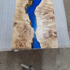 May include: A rectangular wooden table top with a river of blue epoxy resin running through the center. The wood has a light brown color with dark brown knots and patterns. The table top measures approximately 30 inches long.