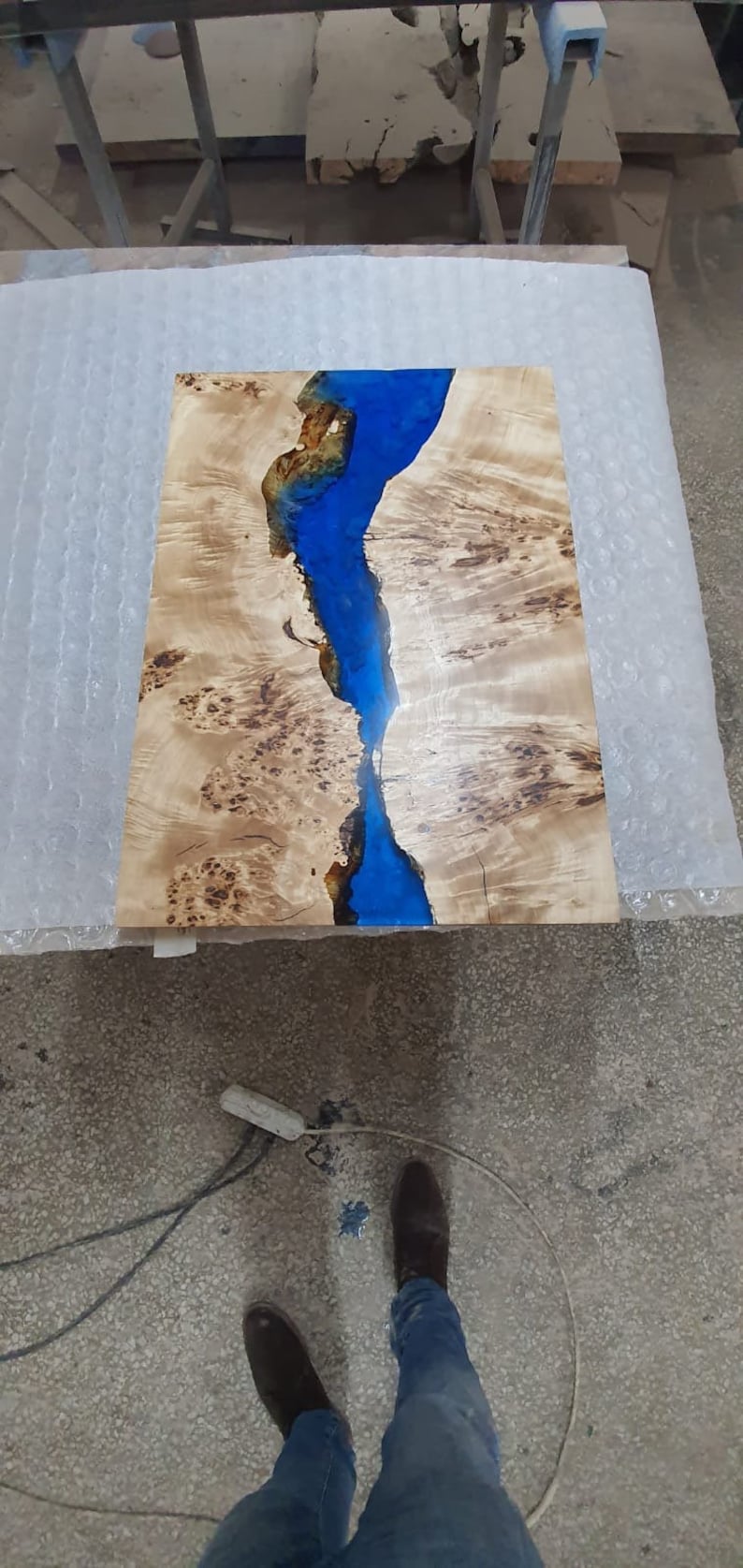 May include: A rectangular wooden table top with a river of blue epoxy resin running through the center. The wood has a light brown color with dark brown knots and patterns. The table top measures approximately 30 inches long.