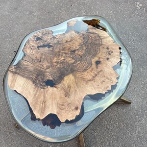 May include: A unique coffee table with a live edge wood slab encased in clear resin. The wood features a rich, dark brown grain pattern. The table is supported by three gold-colored metal legs. The overall dimensions are approximately 36 inches long by 24 inches wide.
