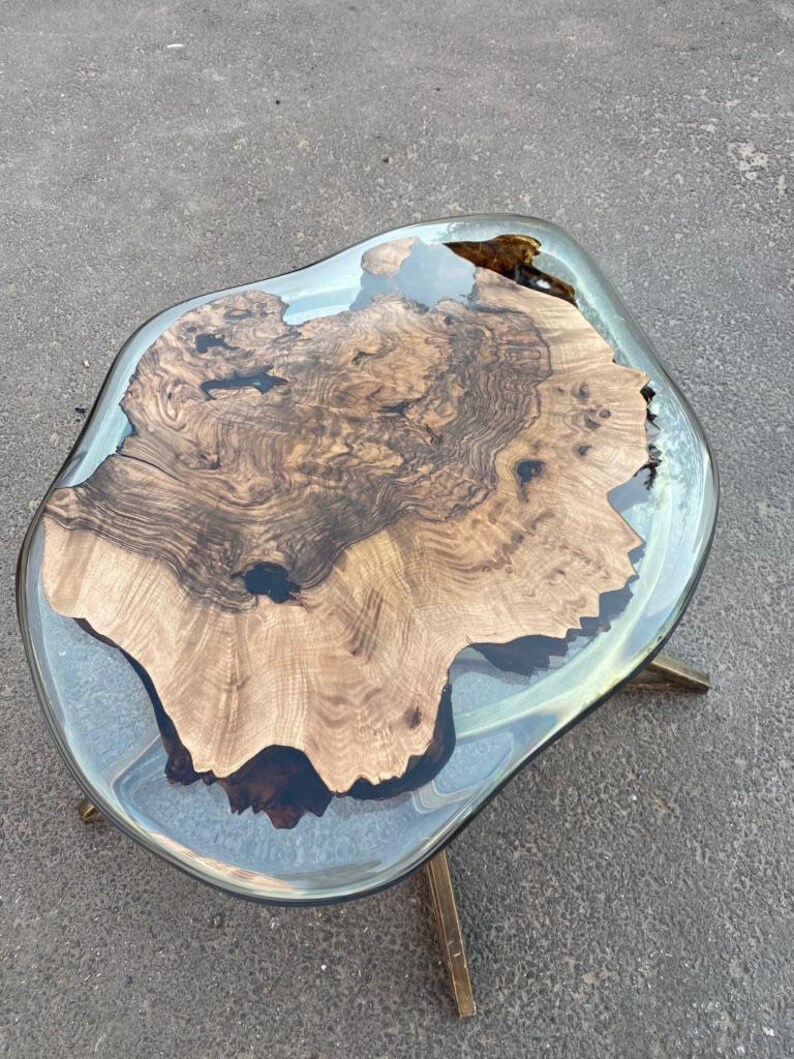 May include: A unique coffee table with a live edge wood slab encased in clear resin. The wood features a rich, dark brown grain pattern. The table is supported by three gold-colored metal legs. The overall dimensions are approximately 36 inches long by 24 inches wide.