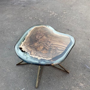 May include: A unique, handcrafted coffee table with a live edge wood top encased in clear resin. The table has a natural wood grain pattern and sits on a gold-colored metal base. The table is approximately 24 inches long, 18 inches wide, and 16 inches high.