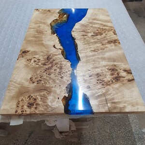 May include: A rectangular wooden table with a river of blue epoxy resin running through the center. The wood has a light brown color with dark brown knots and patterns. The epoxy resin is a vibrant blue.
