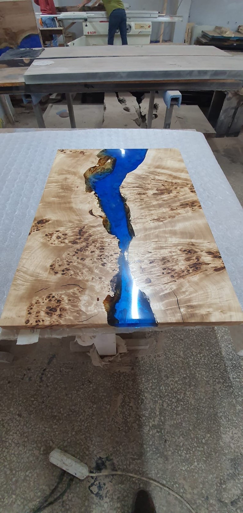 May include: A rectangular wooden table with a river of blue epoxy resin running through the center. The wood has a light brown color with dark brown knots and patterns. The epoxy resin is a vibrant blue.
