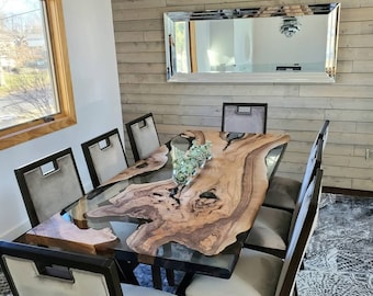 May include: A modern dining table with a live edge wood top and a clear epoxy resin fill. The table is surrounded by six upholstered chairs with dark wood frames. A large mirror hangs on the wood-paneled wall.