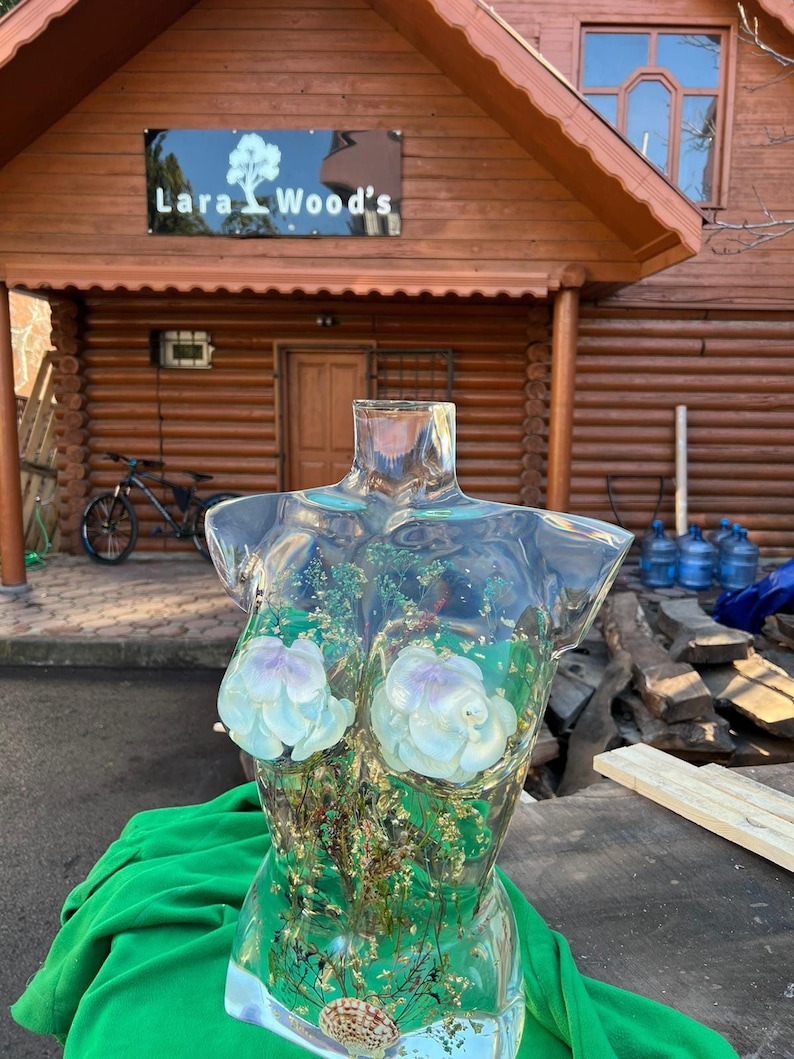 Puede incluir: Una escultura de torso de resina transparente con acentos de flores blancas y moradas, follaje seco y una concha. La escultura se exhibe sobre una tela verde, con un edificio de madera y un letrero que dice "Lara Wood's" en el fondo.