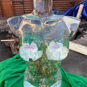 Puede incluir: Una escultura de busto de resina transparente con inclusiones florales y hojas de oro. El busto presenta dos acentos florales blancos y lavanda y una concha en la base. La escultura se exhibe sobre una tela verde.