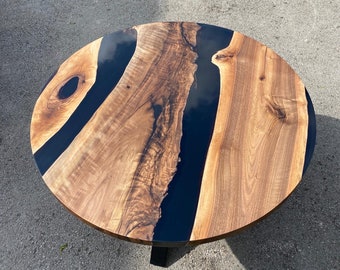 May include: Round coffee table with a live edge walnut wood top and a black epoxy resin river. The table has a dark finish and is supported by a black metal base. The wood grain is visible, adding a natural element to the design.
