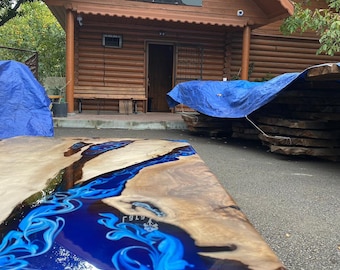 May include: A handcrafted wooden table with a blue resin river design. The table features a natural wood grain and a vibrant blue epoxy resin, creating a unique and artistic piece. The table is set outside a log cabin.