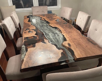 May include: A large, rectangular wooden dining table with a river-like design in the center. The table is made of natural wood with a dark, glossy epoxy resin. The table is surrounded by eight upholstered chairs.