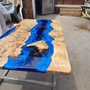 Op de afbeelding: Een rechthoekige houten tafel met een blauw epoxyhars rivierontwerp. De tafel heeft een natuurlijke houtnerf afwerking met een glanzende blauwe harsrivier. De tafel wordt ondersteund door een metalen frame. Ongeveer 180 cm lang.