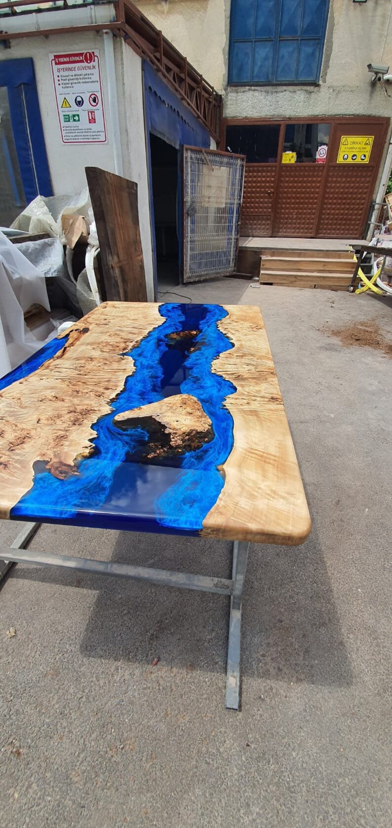 Op de afbeelding: Een rechthoekige houten tafel met een blauw epoxyhars rivierontwerp. De tafel heeft een natuurlijke houtnerf afwerking met een glanzende blauwe harsrivier. De tafel wordt ondersteund door een metalen frame. Ongeveer 180 cm lang.
