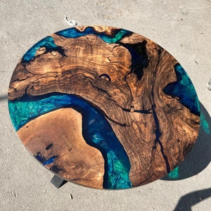 Op de afbeelding: Ronde houten salontafel met een uniek ontwerp. De tafel heeft een natuurlijk houtnerfpatroon met een rivier van blauwe en turquoise epoxyhars. De tafel wordt ondersteund door een zwarte metalen basis. Diameter ongeveer 100 cm.