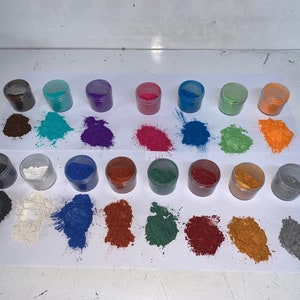 Pode incluir: Uma cole&ccedil;&atilde;o de pequenos frascos transparentes cheios de p&oacute;s coloridos. Os p&oacute;s est&atilde;o espalhados em papel branco, mostrando um espectro de tons, incluindo marrom, turquesa, roxo, rosa, azul, verde e laranja. A imagem mostra uma variedade de pigmentos.
