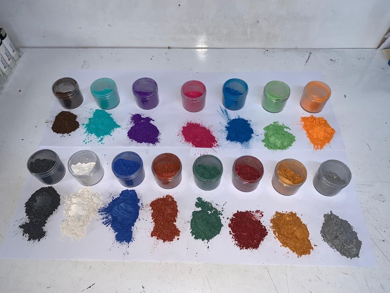 Pode incluir: Uma cole&ccedil;&atilde;o de pequenos frascos transparentes cheios de p&oacute;s coloridos. Os p&oacute;s est&atilde;o espalhados em papel branco, mostrando um espectro de tons, incluindo marrom, turquesa, roxo, rosa, azul, verde e laranja. A imagem mostra uma variedade de pigmentos.