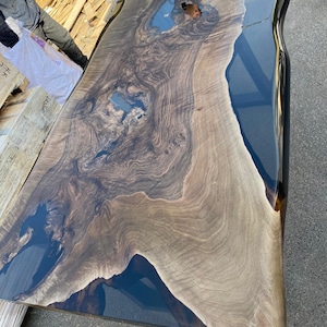 May include: A large, live edge wooden table with a dark blue epoxy resin river. The wood grain is a mix of light and dark brown, creating a natural, organic pattern. The table has a smooth, glossy finish and is likely a custom piece.