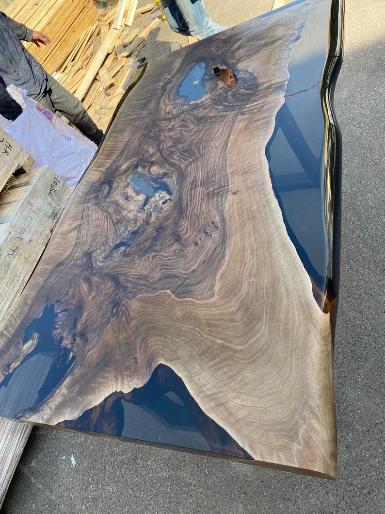 May include: A large, live edge wooden table with a dark blue epoxy resin river. The wood grain is a mix of light and dark brown, creating a natural, organic pattern. The table has a smooth, glossy finish and is likely a custom piece.