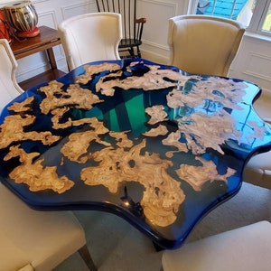 May include: A unique, custom-made epoxy resin table with a striking design. The table features a deep blue resin base with natural wood elements, creating a flowing, organic aesthetic. The table is surrounded by cream-colored chairs.