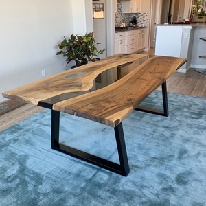 May include: A large, custom-made wooden dining table with a live edge design. The table features a dark, clear epoxy resin river running down the center. The wood has a natural, light brown color. The table is supported by black metal legs.