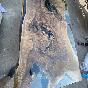May include: A large, rectangular wooden table with a live edge and a glossy, dark finish. The wood grain is prominent, with areas of dark, almost black, resin filling natural voids. The table's surface reflects the sky, creating a striking visual effect.