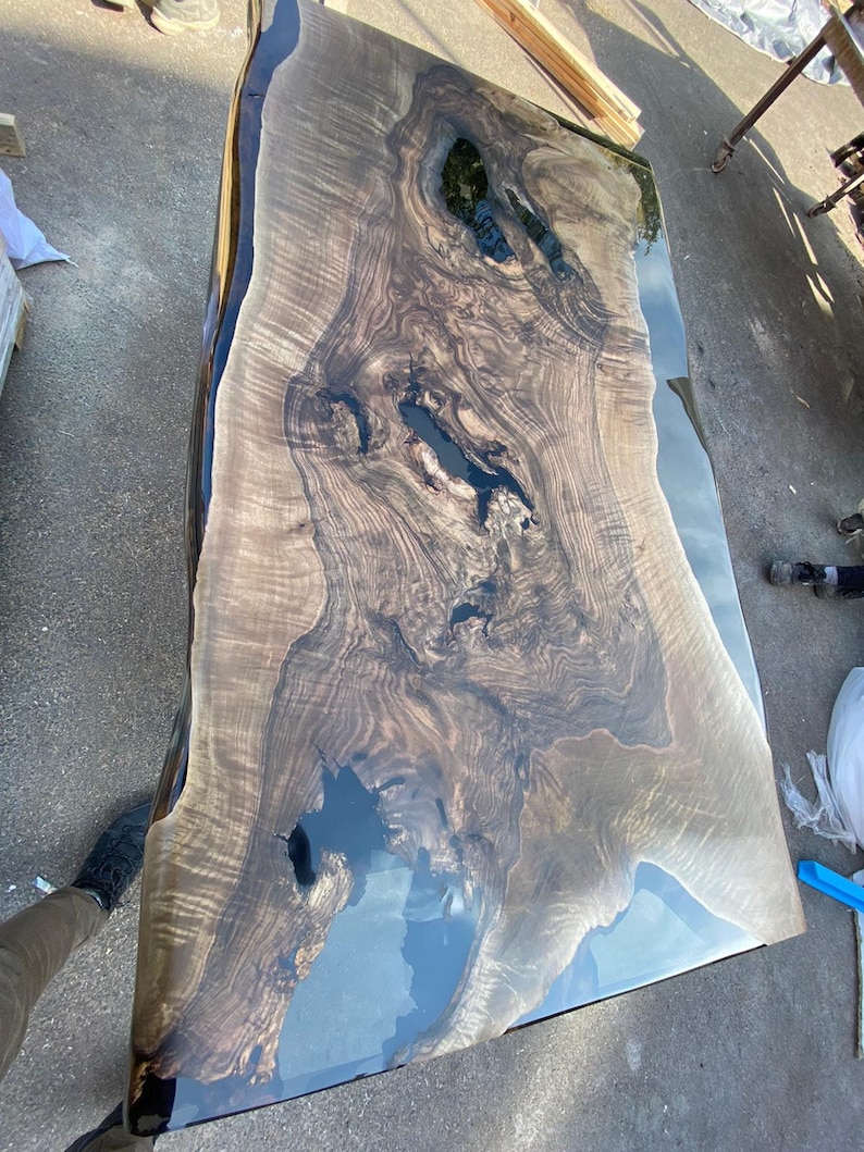 May include: A large, rectangular wooden table with a live edge and a glossy, dark finish. The wood grain is prominent, with areas of dark, almost black, resin filling natural voids. The table's surface reflects the sky, creating a striking visual effect.