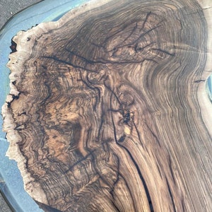 May include: A close-up view of a wooden table with a clear resin border. The table's surface showcases a rich, dark brown wood grain pattern with intricate details. The resin edge adds a modern touch to the natural wood.