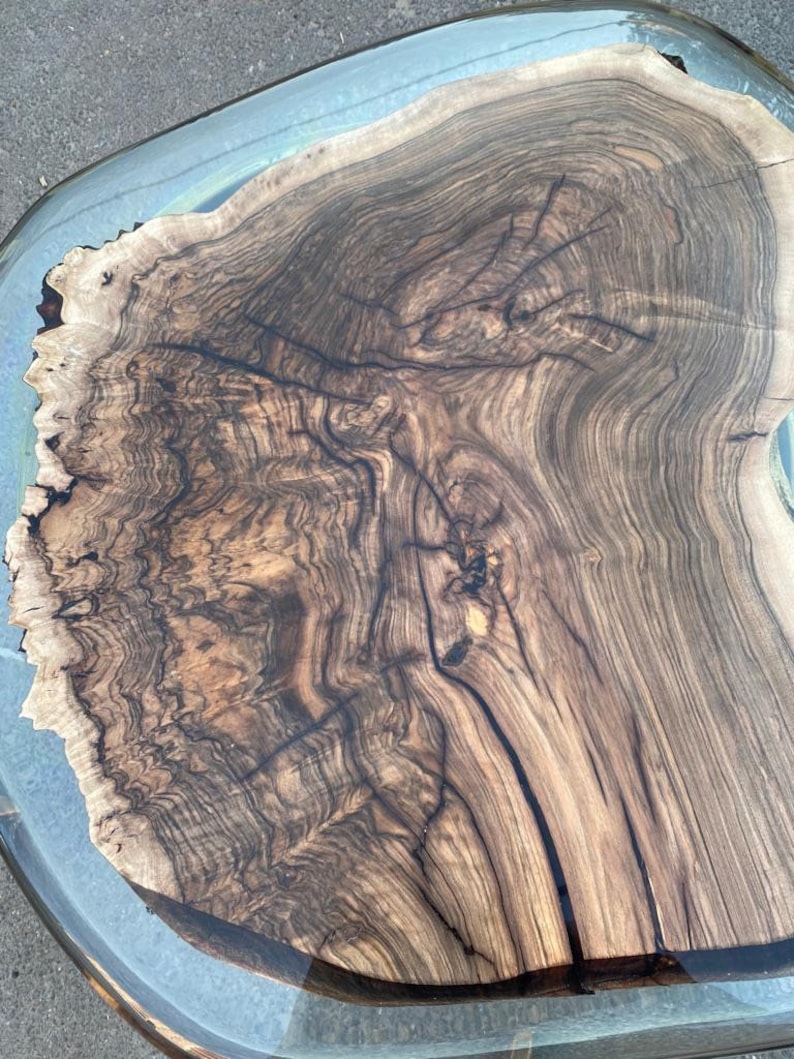 May include: A close-up view of a wooden table with a clear resin border. The table's surface showcases a rich, dark brown wood grain pattern with intricate details. The resin edge adds a modern touch to the natural wood.