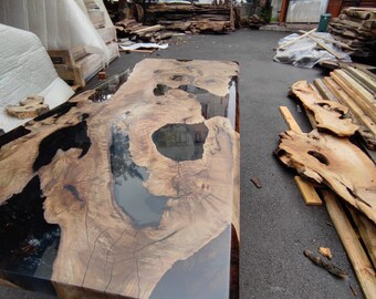 Puede incluir: Una mesa rectangular de madera con una superficie oscura y brillante. La mesa presenta un patrón de vetas de madera natural con áreas de resina negra. La mesa está al aire libre, con un edificio y tablas de madera en el fondo.