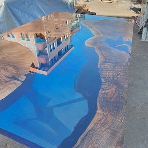 Könnte beinhalten: Großer, rechteckiger Holztisch mit einem blauen Epoxidharzfluss. Das Holz hat eine dunkle, natürliche Maserung. Das Harz reflektiert den Himmel und ein Gebäude. Der Tisch befindet sich im Freien.