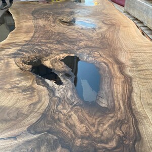 May include: A large, rectangular wooden table with a natural, organic design. The table features a rich, dark brown wood grain with lighter brown and black accents. The surface has a glossy finish and includes a dark, resin-filled void.