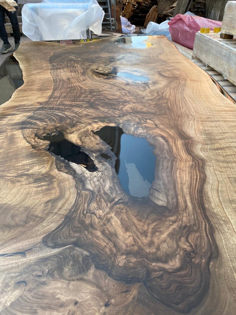 May include: A large, rectangular wooden table with a natural, organic design. The table features a rich, dark brown wood grain with lighter brown and black accents. The surface has a glossy finish and includes a dark, resin-filled void.