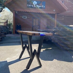 May include: A handcrafted wooden table with a dark wood top and blue resin accents. The table has black metal legs with a unique zigzag design. The business name "Lara Wood's" is visible in the background.